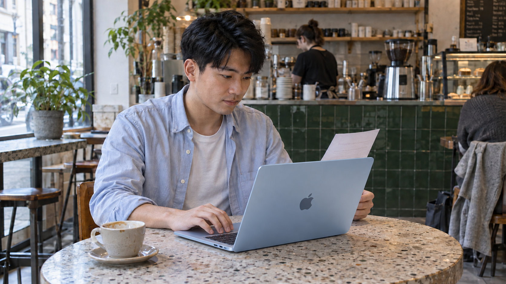 A young man compares a printed document with a sky blue MacBook Air at a cafe table.